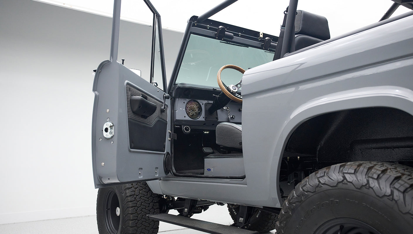 Open door view of gray Classic Ford Bronco with visible steering wheel, dashboard, and seating inside white studio.