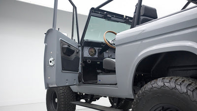 Open door view of gray Classic Ford Bronco with visible steering wheel, dashboard, and seating inside white studio.
