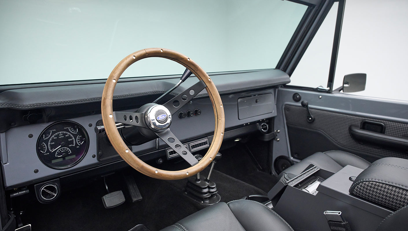 Gray Classic Ford Bronco interior showing wood steering wheel, dashboard, pedals, and center console.
