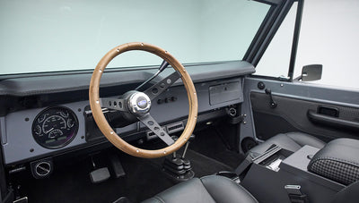 Gray Classic Ford Bronco interior showing wood steering wheel, dashboard, pedals, and center console.