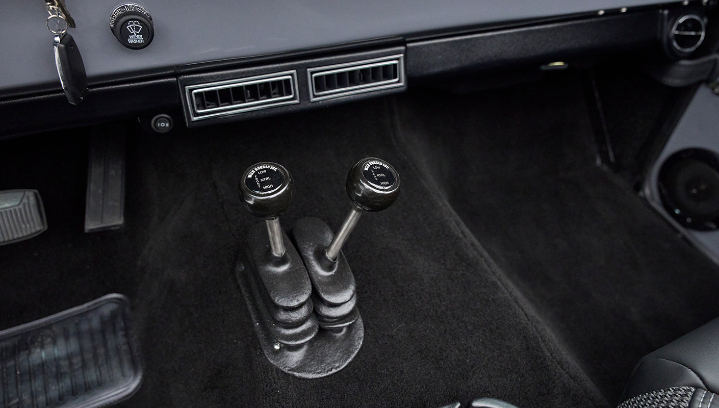 Two black gear shift levers with labeled knobs on black carpet floor of Classic Ford Bronco interior