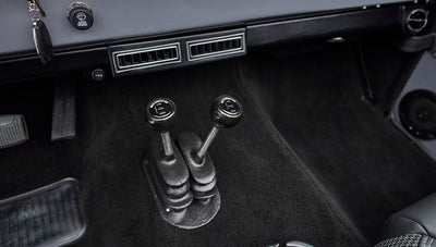 Two black gear shift levers with labeled knobs on black carpet floor of Classic Ford Bronco interior