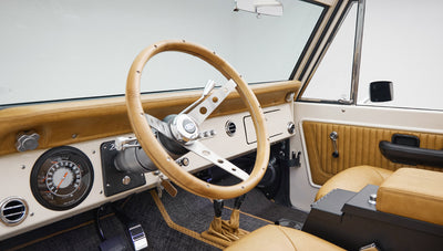Classic Ford Bronco driver cabin with tan leather seats, wood steering wheel, and white dash