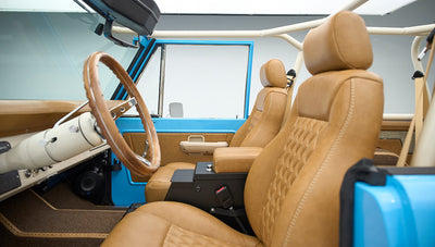 Interior front seats and dashboard of bright blue Classic Ford Bronco with tan upholstery and roll cage