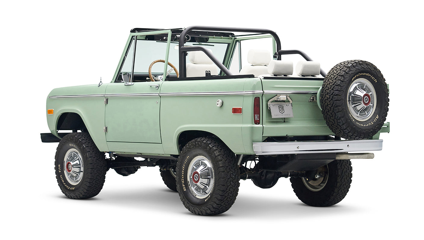 Classic Ford Bronco in pale green with white interior and black roll bar, three quarter rear view showing spare tire