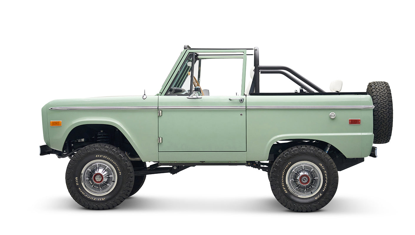 Classic Ford Bronco in pale green with white interior and black roll bar, side view with spare tire on rear