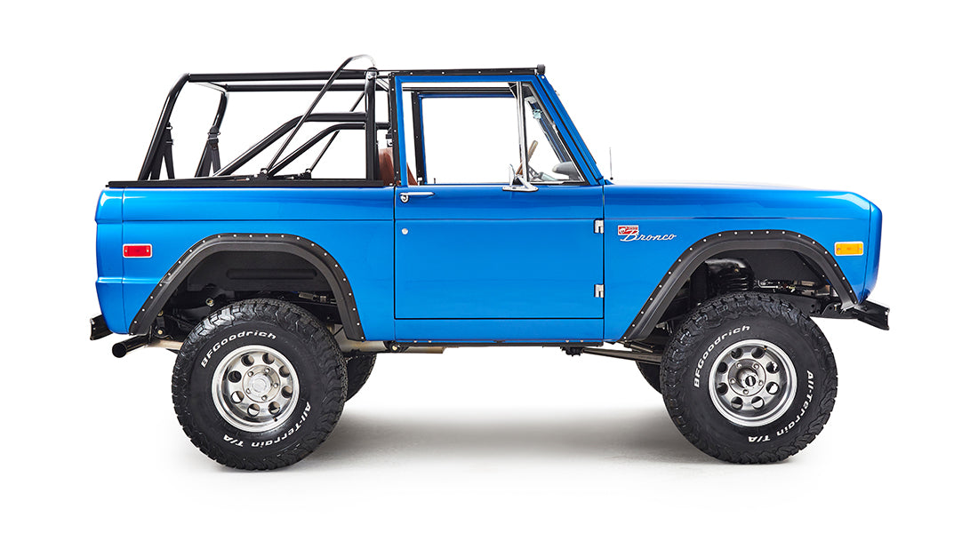 Side view of blue Classic Ford Bronco with black roll cage and off-road tires on white background