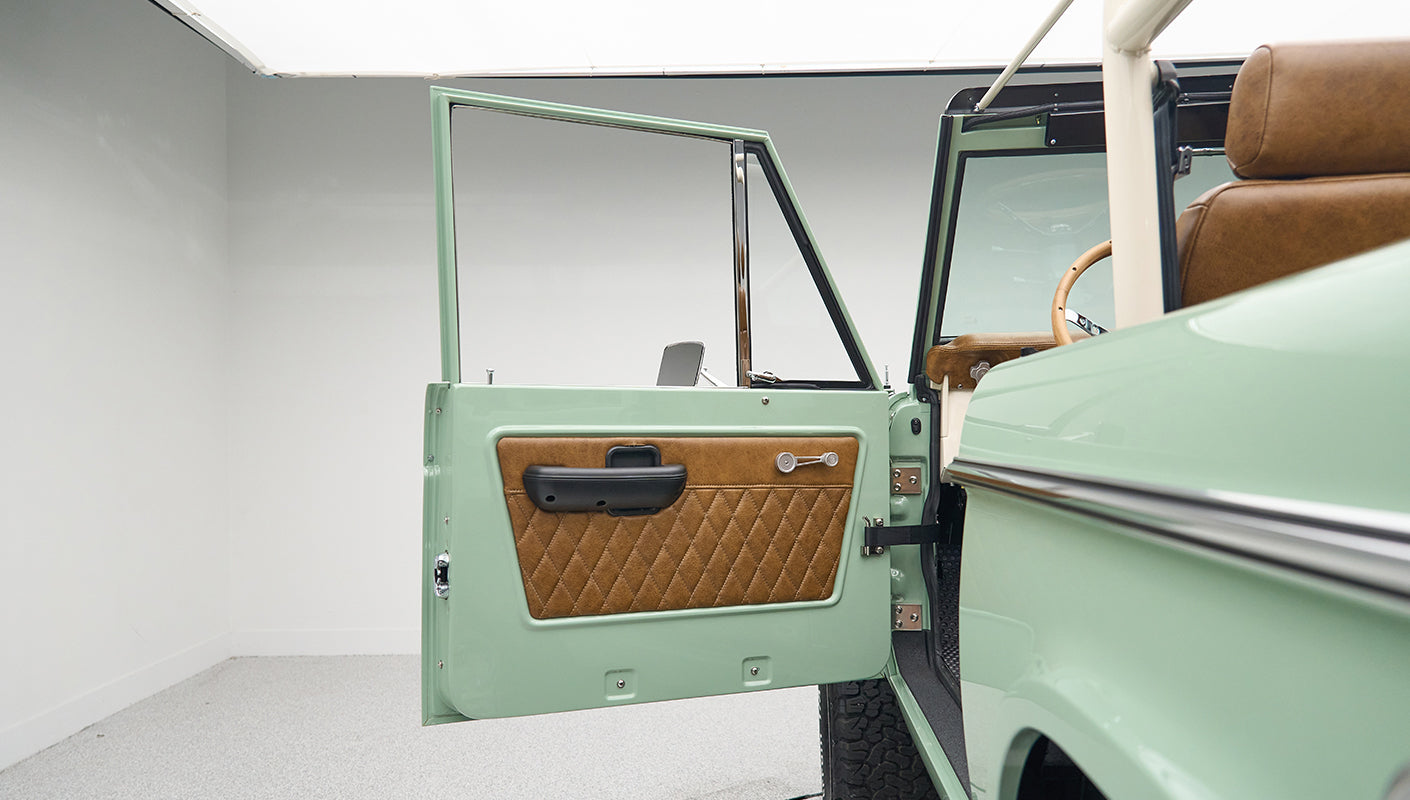 Classic Ford Bronco door open inside a white showroom with green exterior and tan interior upholstery