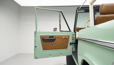 Classic Ford Bronco door open inside a white showroom with green exterior and tan interior upholstery