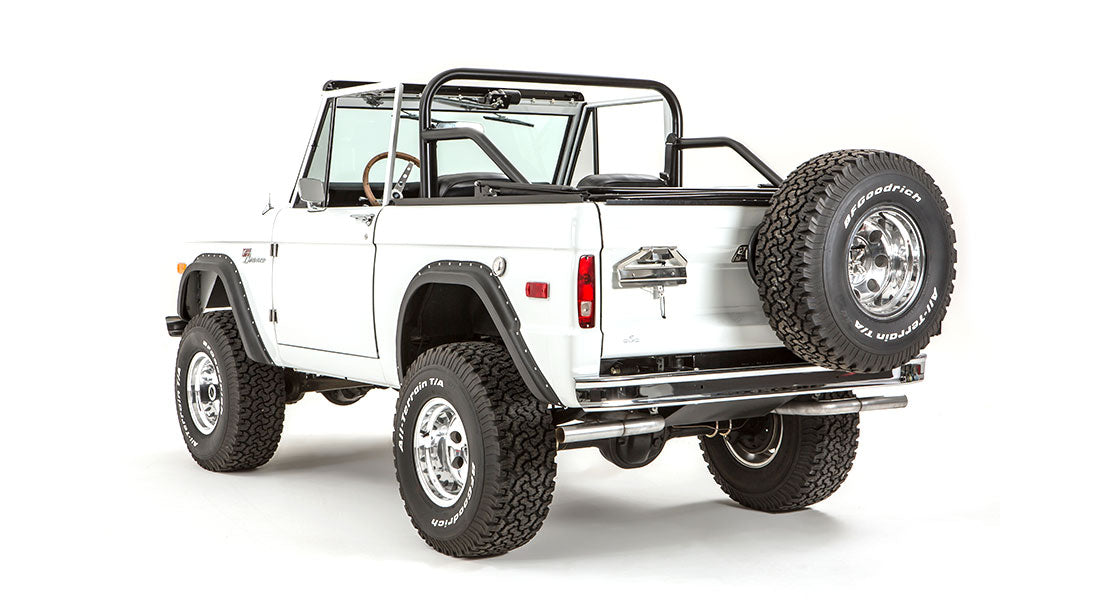 Rear three-quarter view of white Classic Ford Bronco with roll cage, spare tire on tailgate, and black fender flares