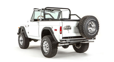 Rear three-quarter view of white Classic Ford Bronco with roll cage, spare tire on tailgate, and black fender flares