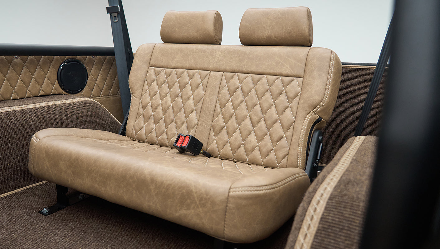 Back bench seat inside Classic Ford Bronco with tan diamond stitching, seat belts, and brown carpeting