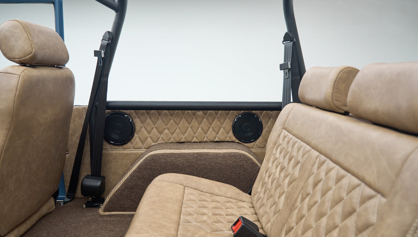 Rear interior of Classic Ford Bronco cabin showing tan diamond-stitched panel with two black speakers and seatbelts