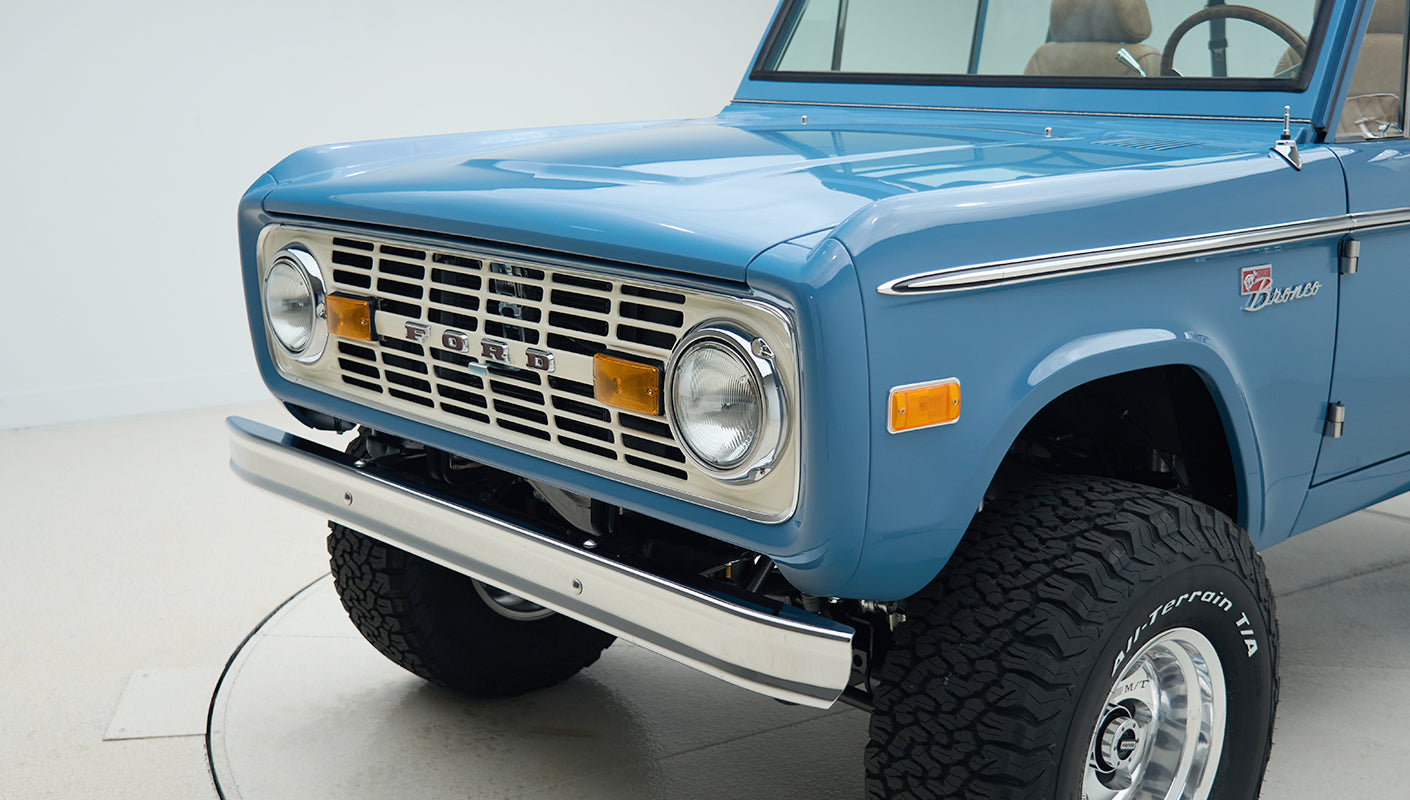 Close-up of front left corner of blue Classic Ford Bronco showing headlights, grille, and bumper
