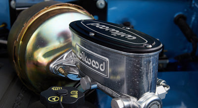 Wilwood brake master cylinder assembly inside engine bay with blue painted panel in background.