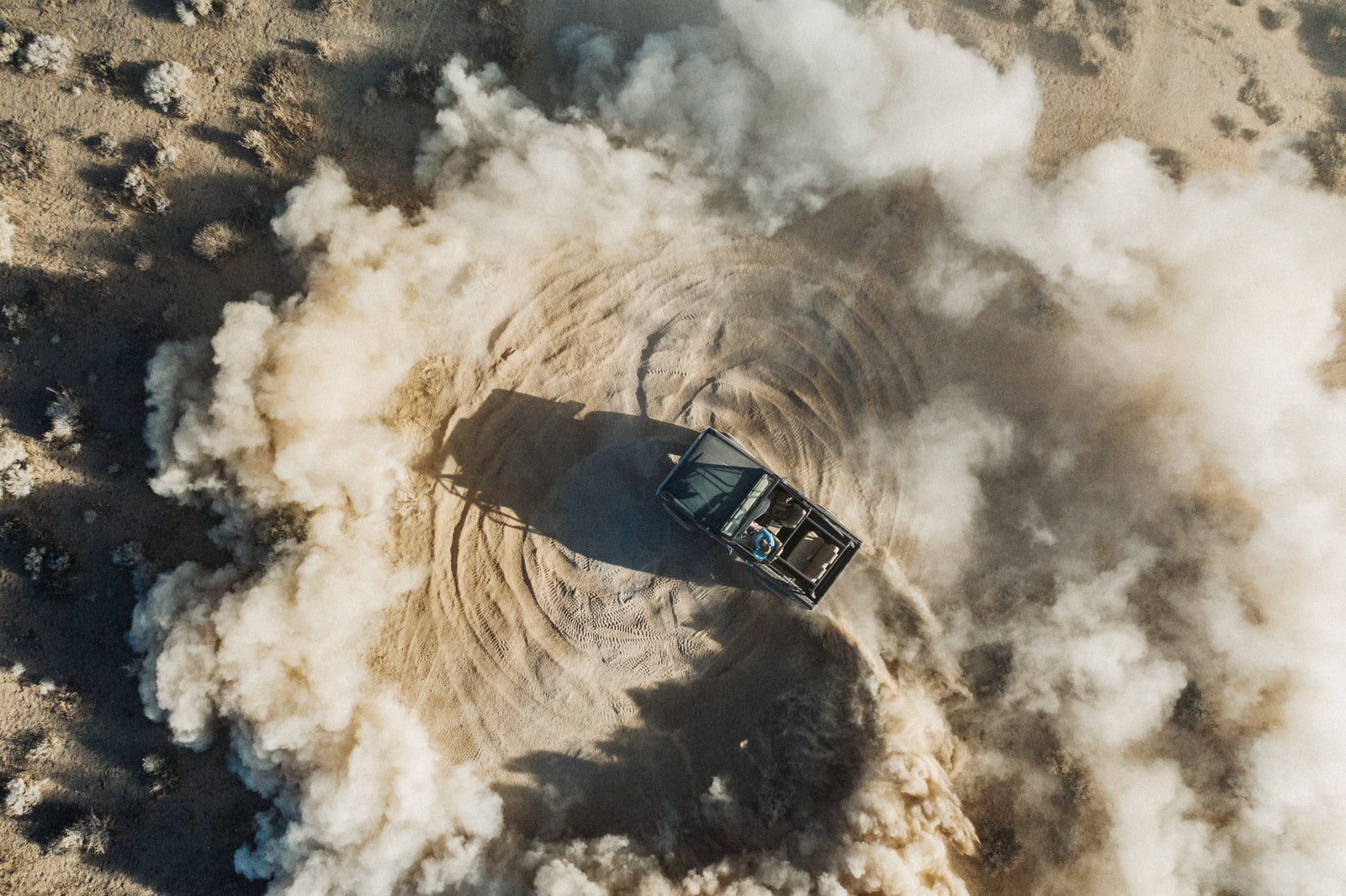 A classic Ford Bronco performing a donut maneuver in a dusty dirt area, aerial view.