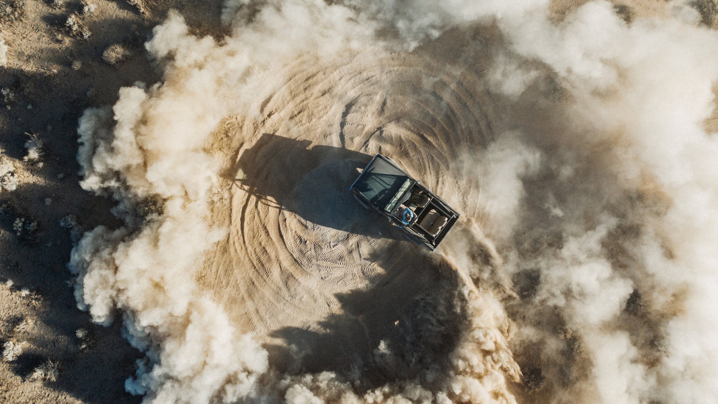 A classic Ford Bronco performing a donut, viewed from above, kicking up dust.