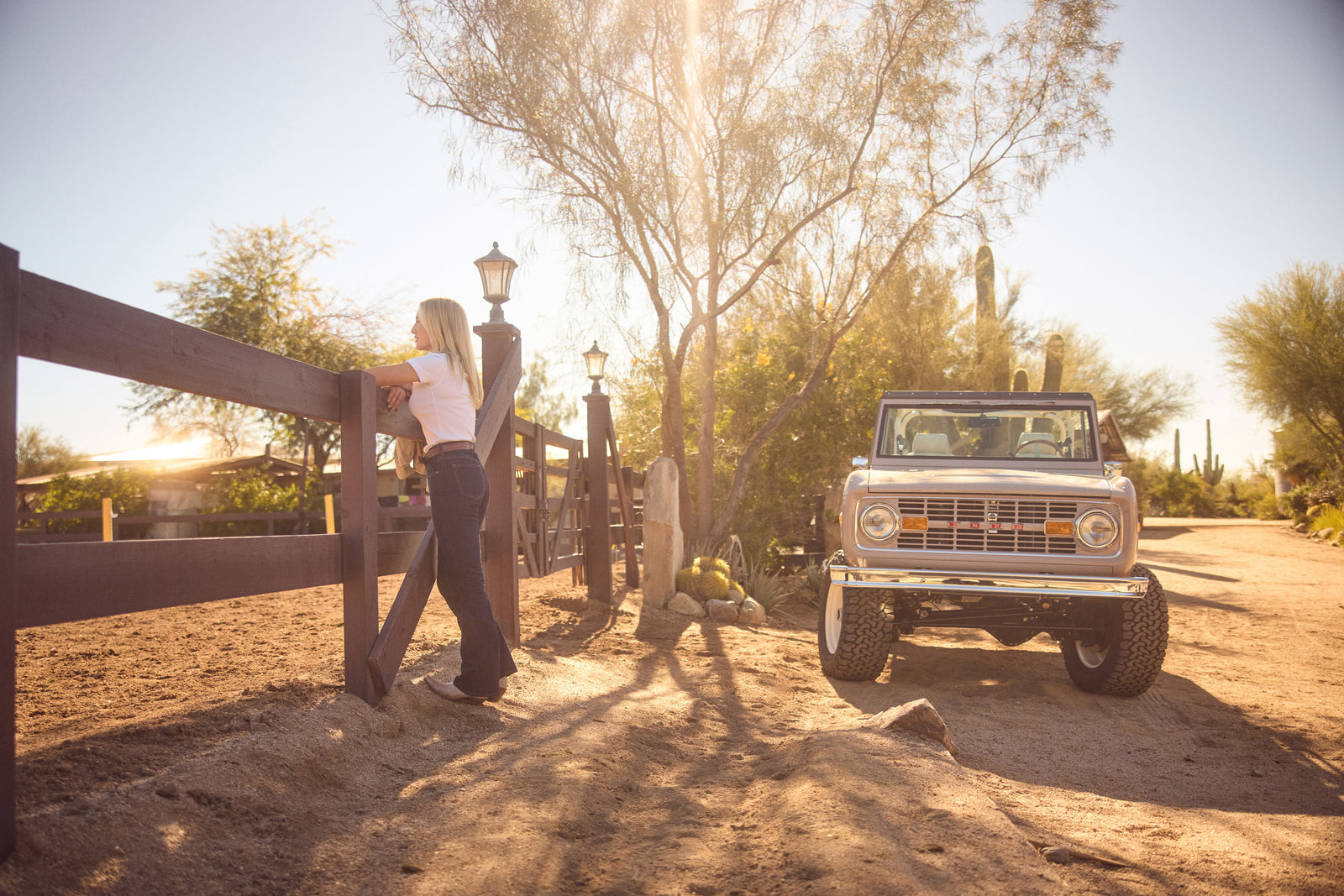Classic Ford Bronco parked on desert dirt road with woman leaning on nearby fence bathed in golden sunlight