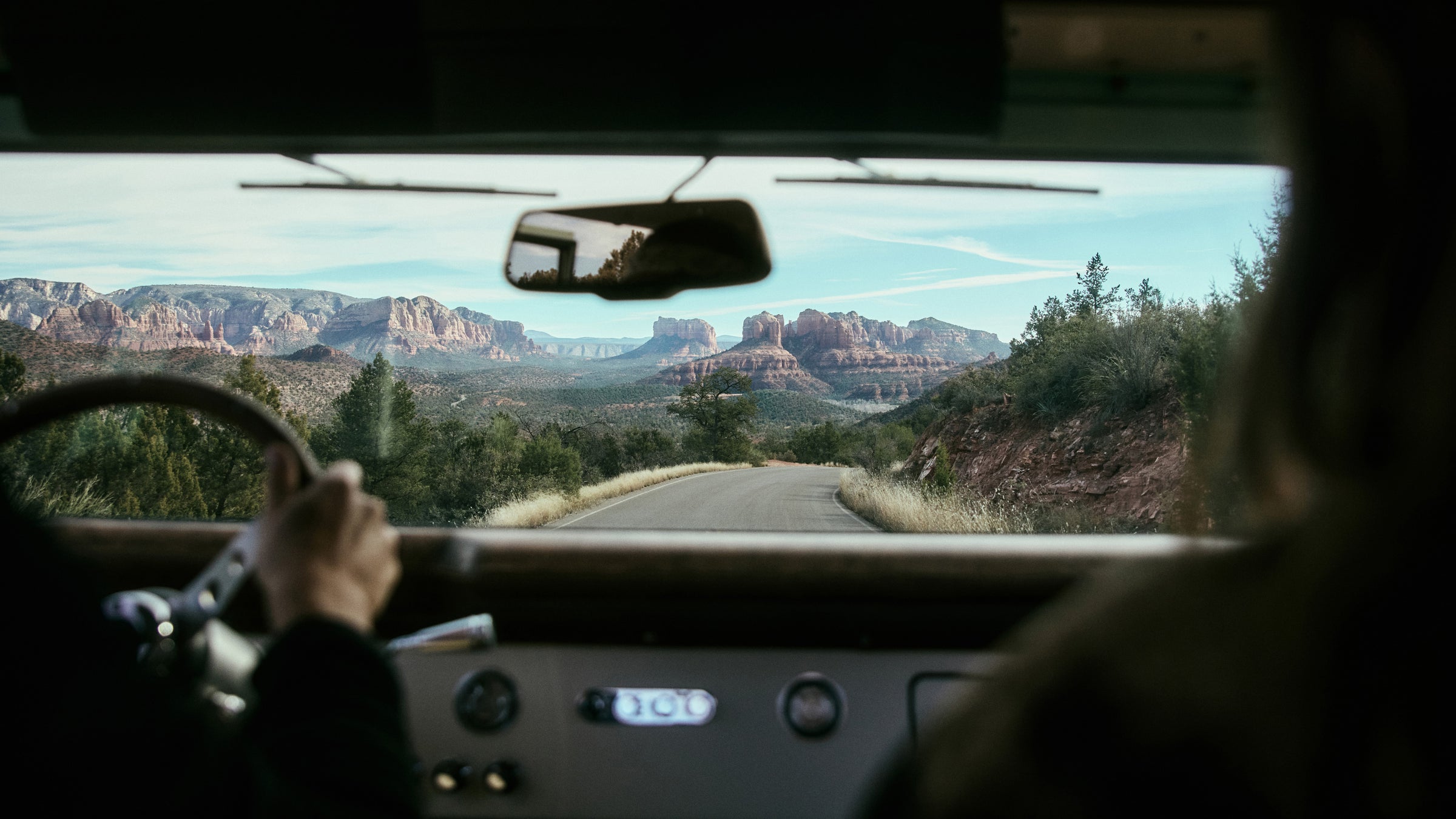 View over steering wheel of person driving Classic Ford Bronco on a winding road with red rock formations in distance