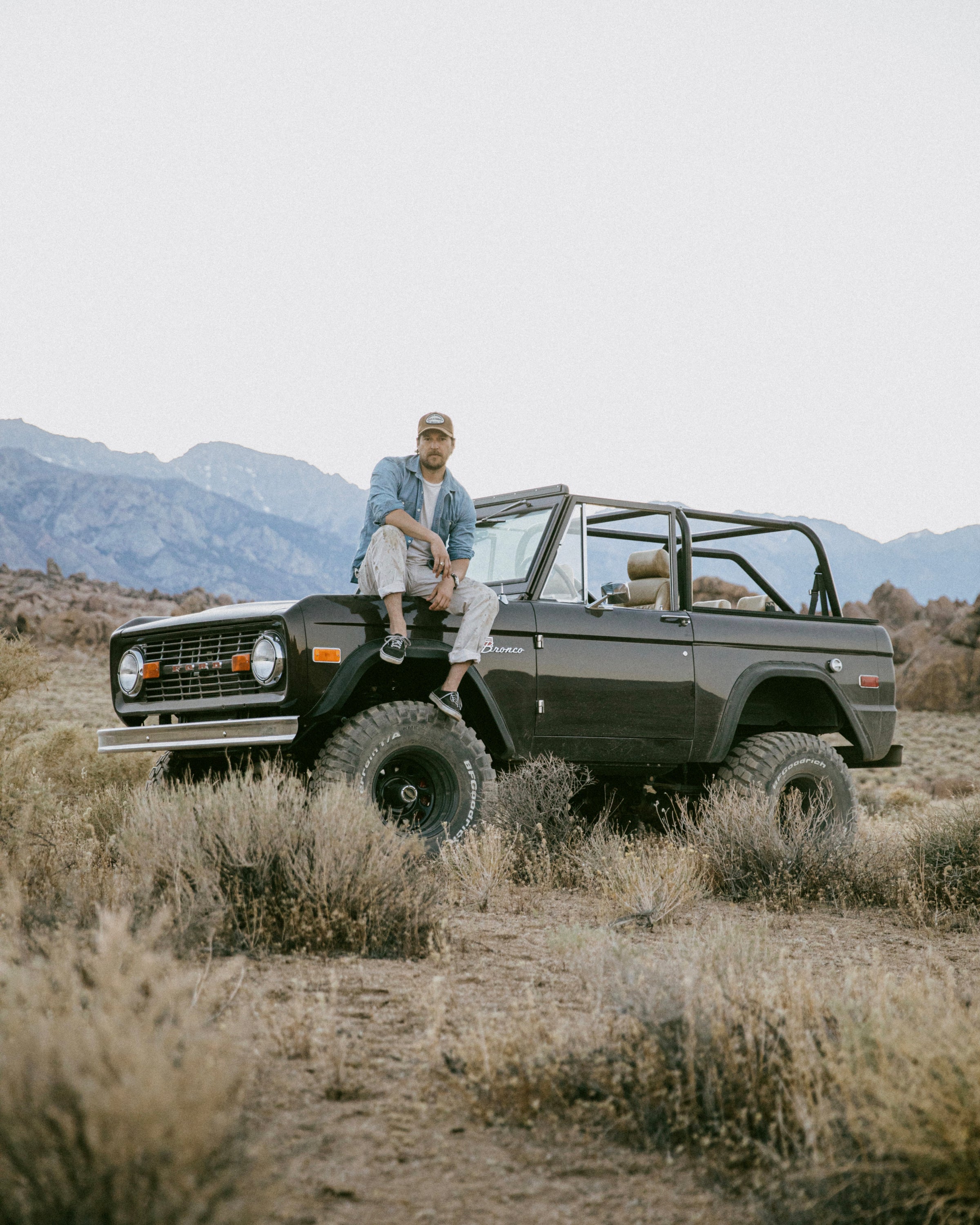 Man sitting on the hood of a black classic Ford Bronco parked in a dry landscape with mountains in the background.