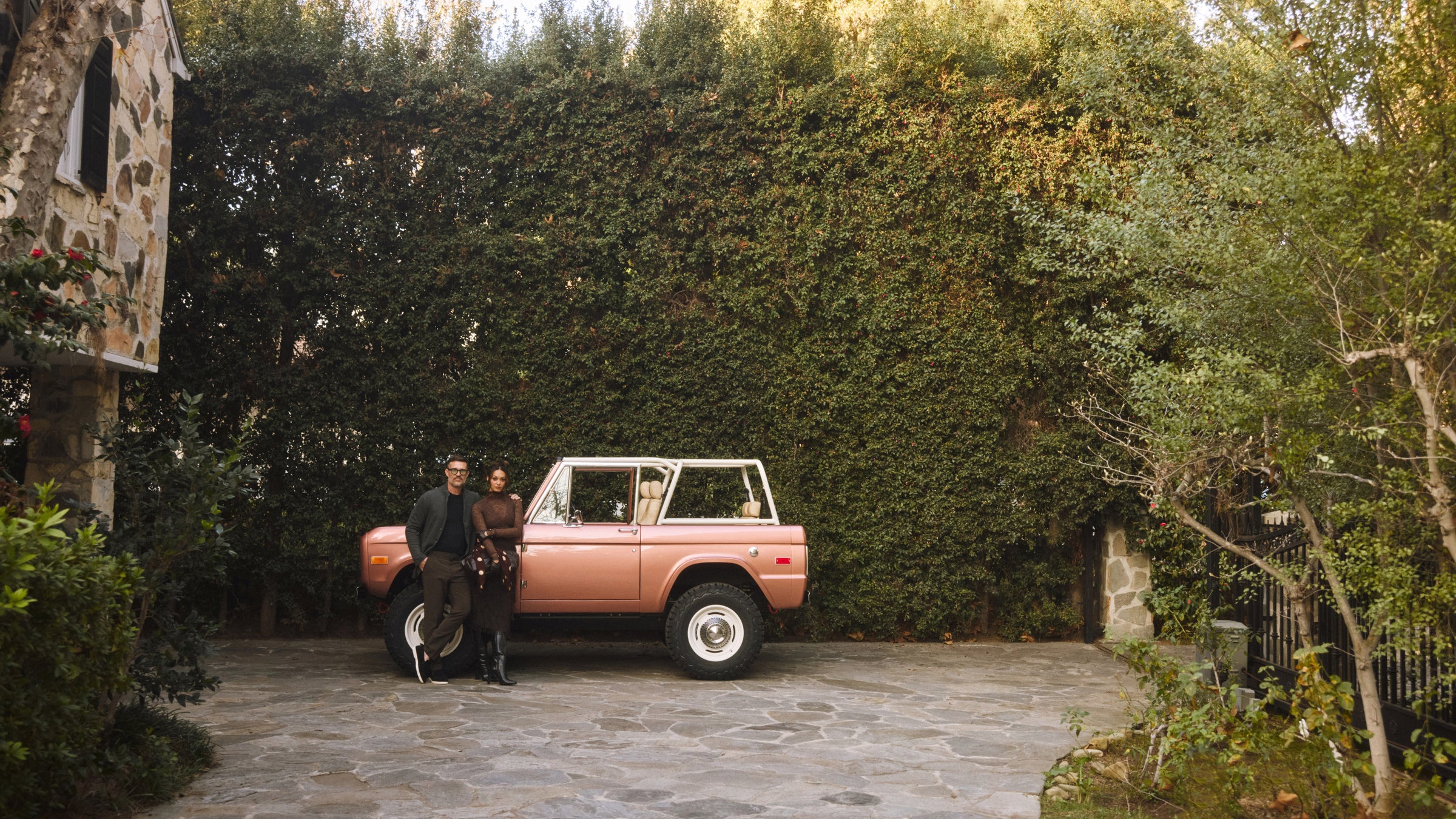 Man and woman leaning on a pink classic Ford Bronco parked on a stone driveway in front of a house and tall greenery.