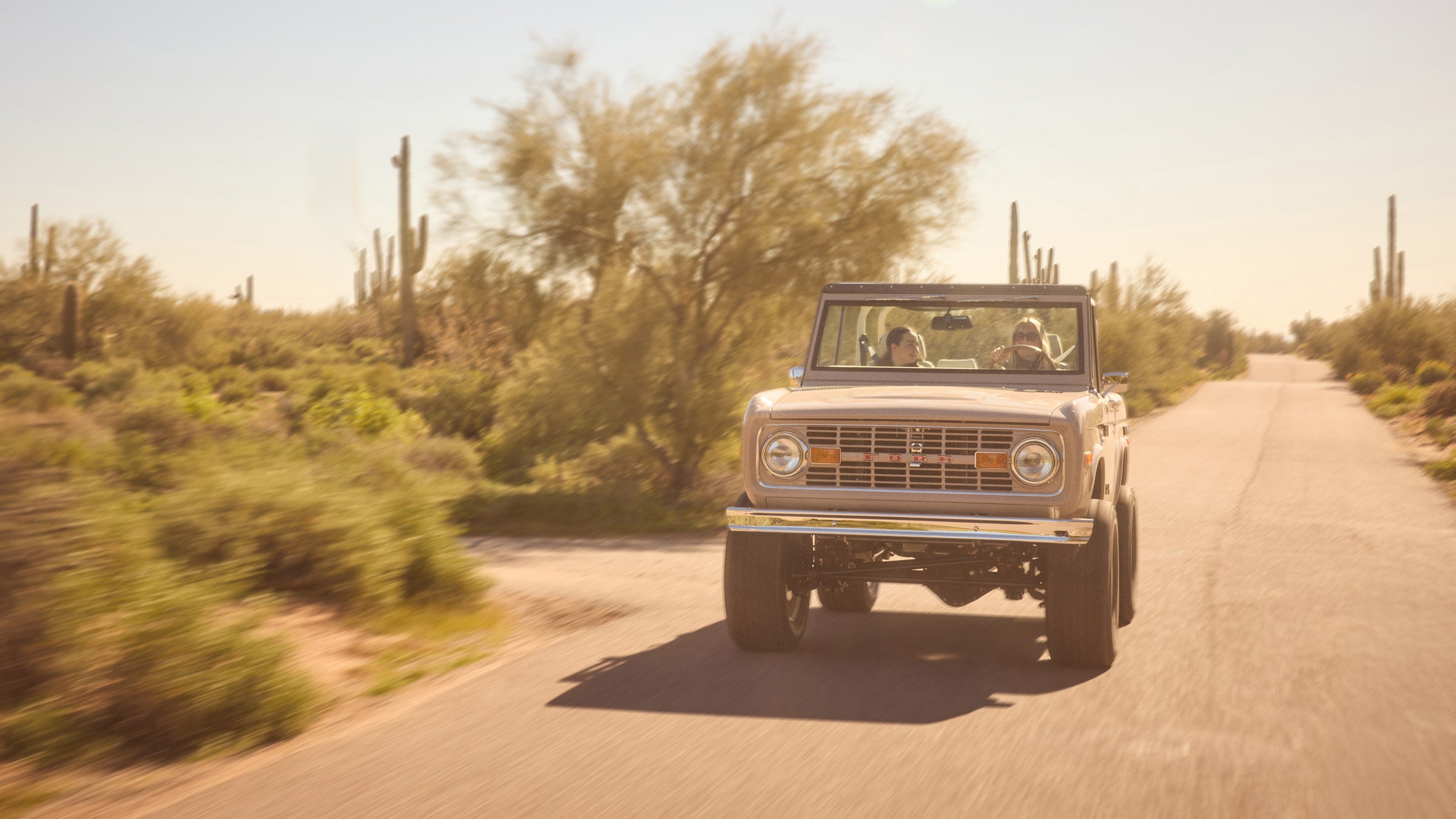 Classic Ford Bronco driving on sunny rural road surrounded by desert plants, two people inside