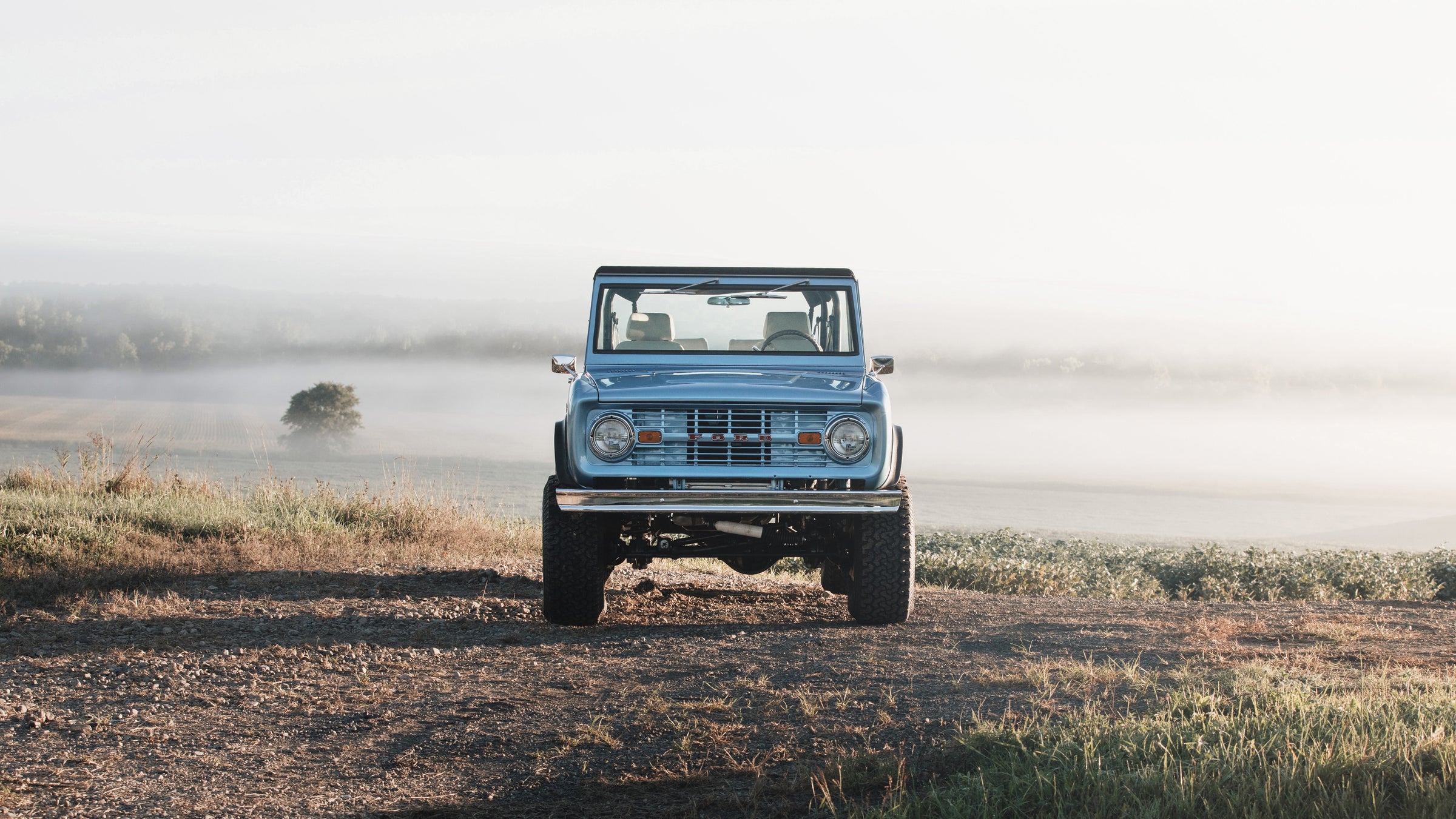 Classic Ford Bronco facing camera on dirt path with foggy countryside and trees in background