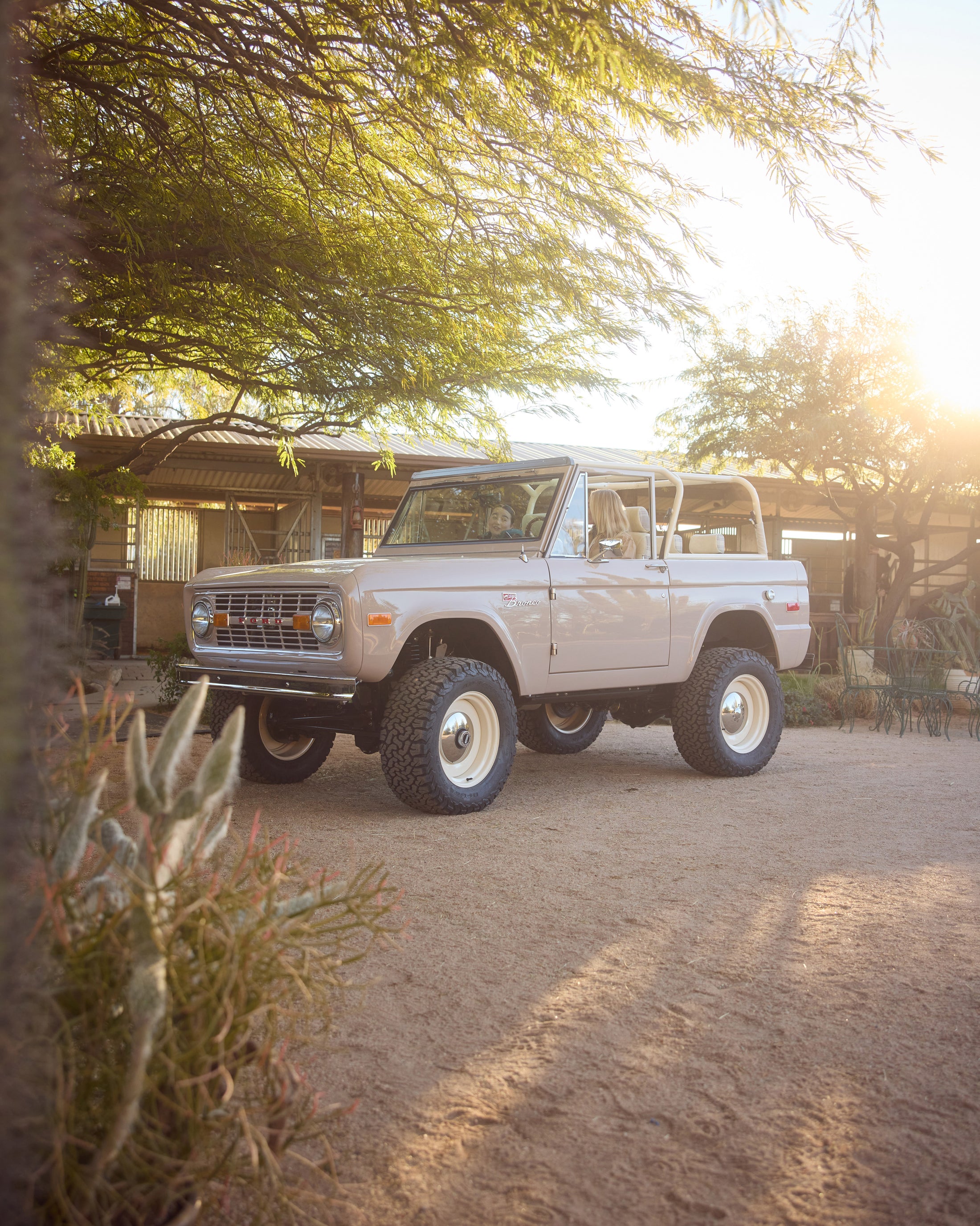 Classic Ford Bronco parked on sunlit dirt surface near farm with two people inside