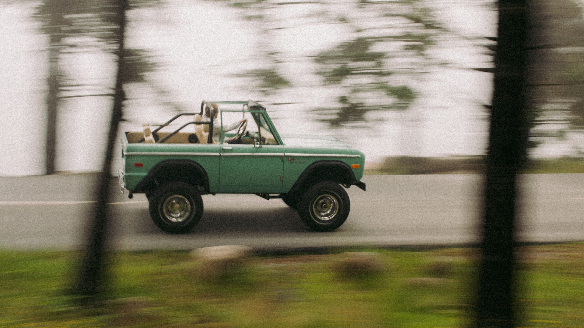 Classic Ford Bronco speeding by on road, side view, motion blurred trees in foreground