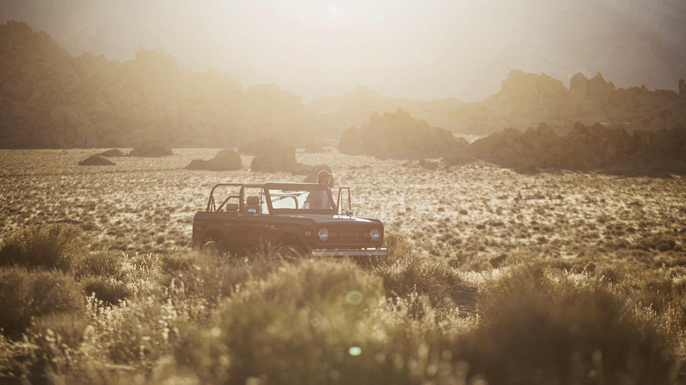 Classic Ford Bronco with person standing inside in a sunlit desert landscape with rocks and brush