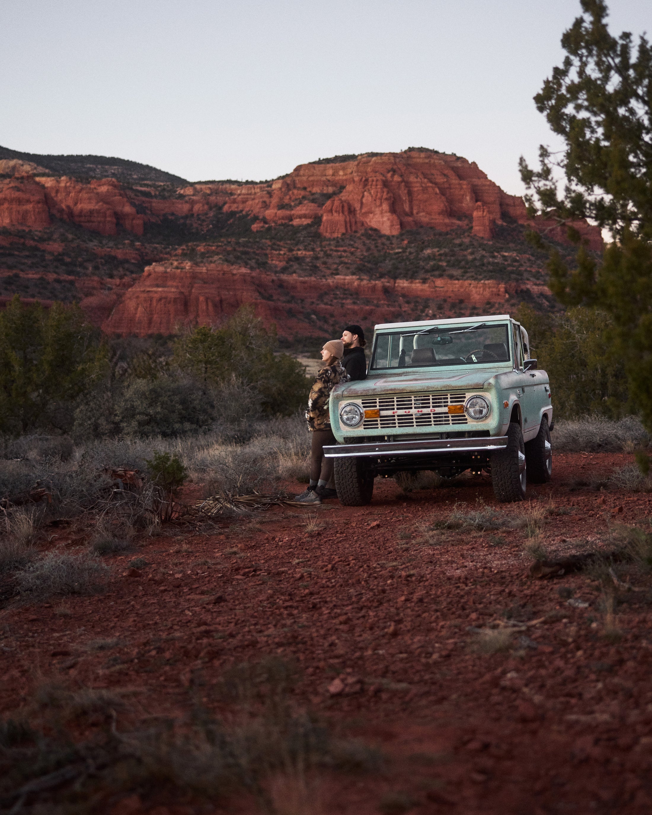 Classic Ford Bronco parked with two people leaning against it in a red rock landscape at dusk