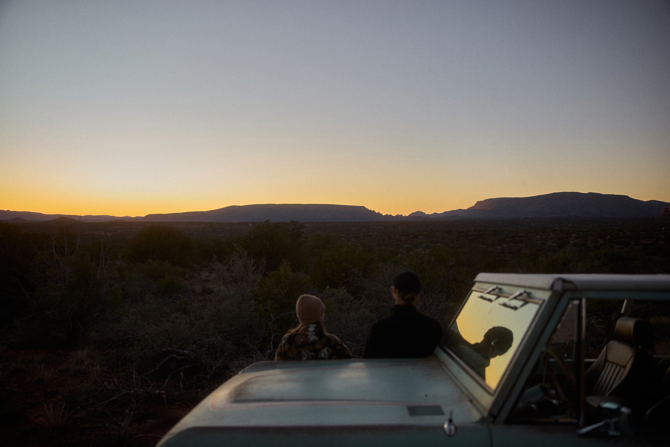 Two people seated on the hood of a vehicle watching a sunset over a desert landscape