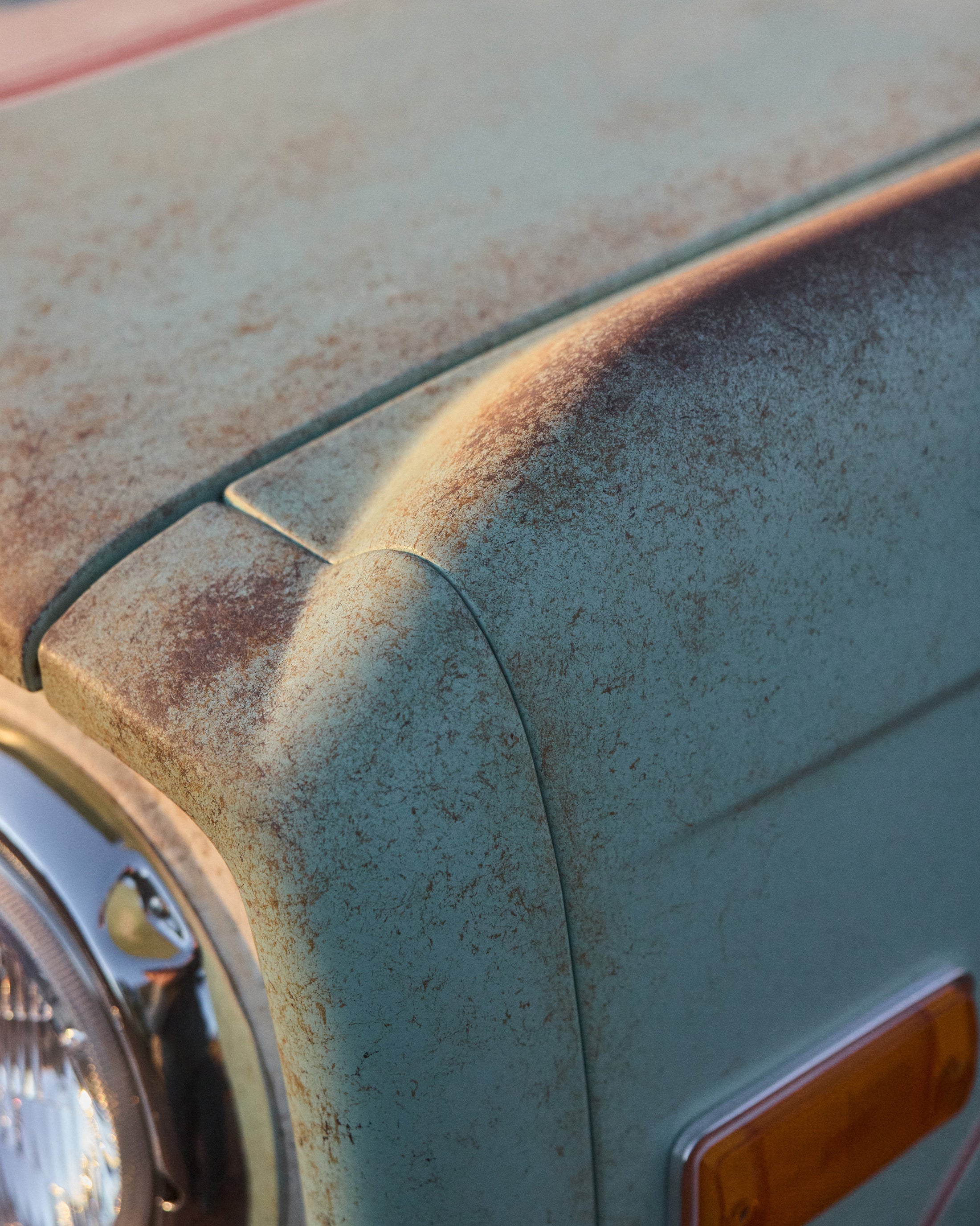 Close-up of a faded and weathered light blue classic vehicle hood with rust spots near a headlight and amber side marker.