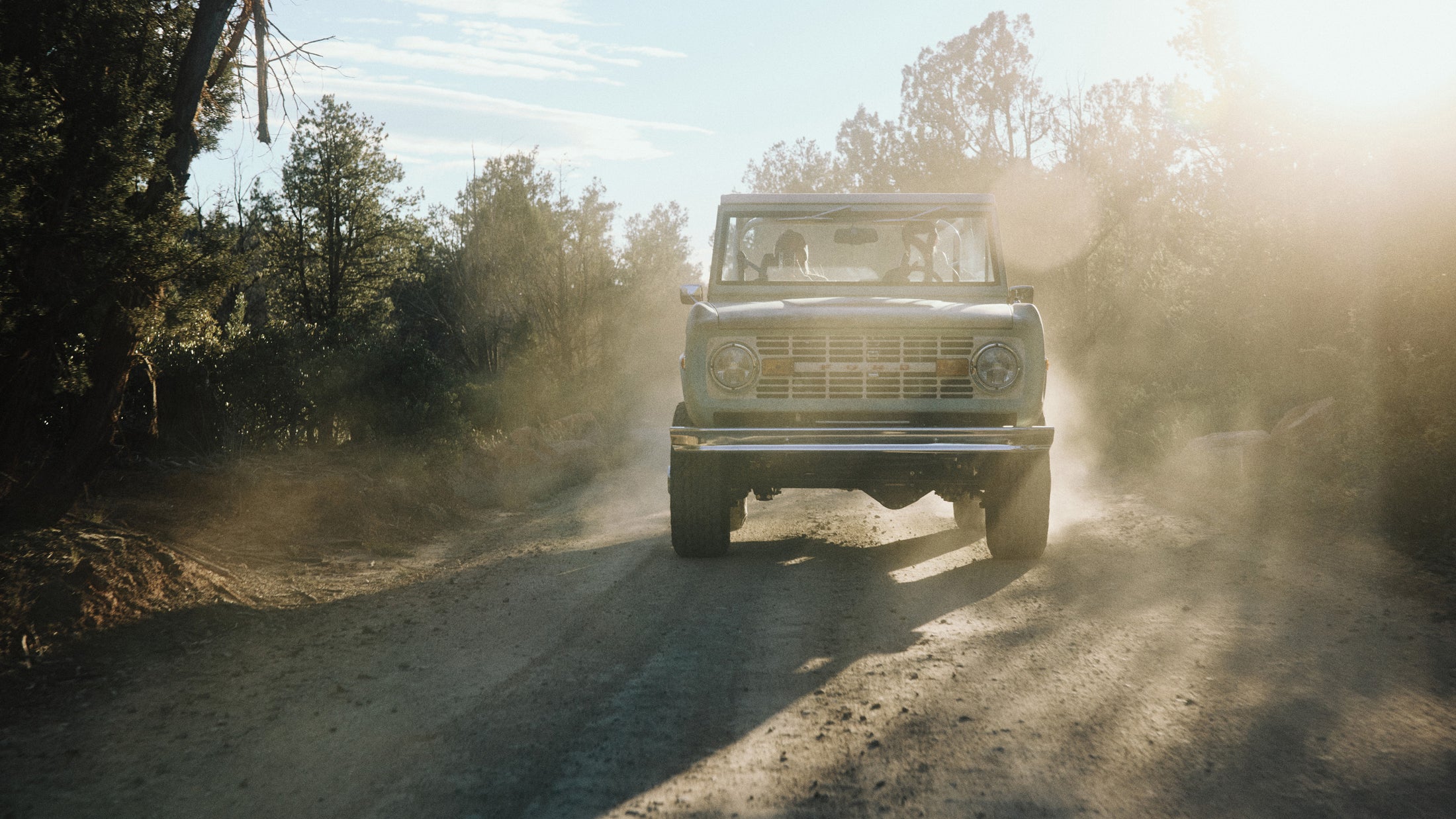 Classic Ford Bronco driving on a dirt road kicking up dust with sunlight filtering through trees