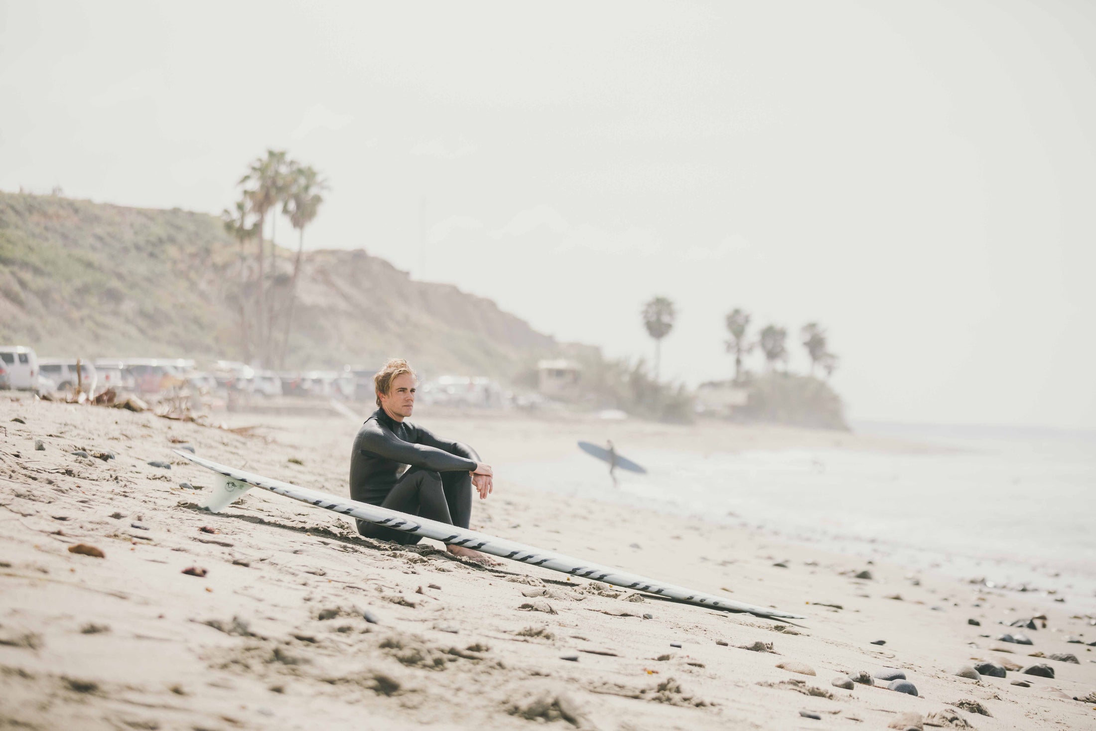 Man in wetsuit sitting on sandy beach beside surfboard with hills and palm trees in background