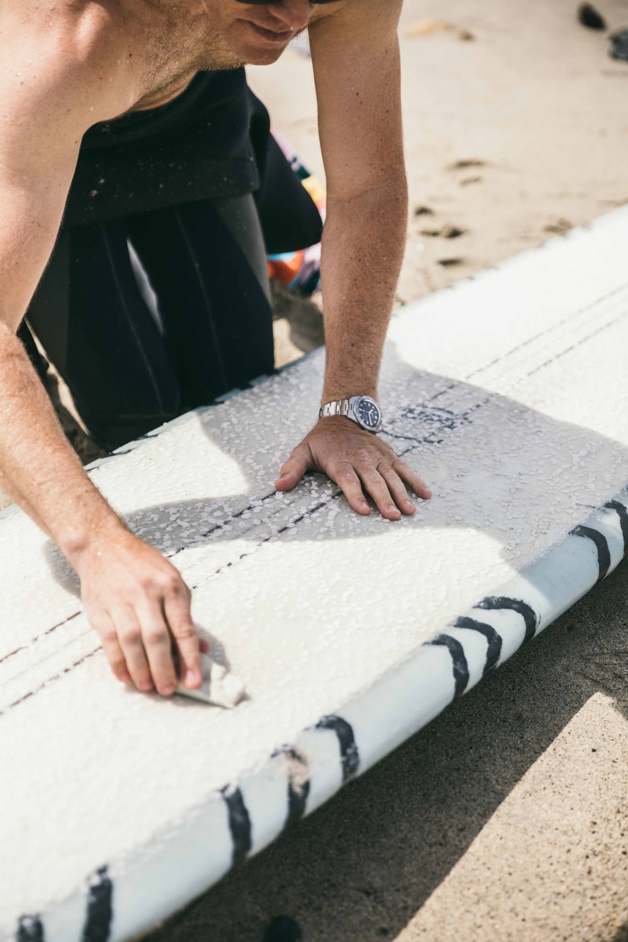 Close-up of man waxing surfboard on sandy beach wearing watch