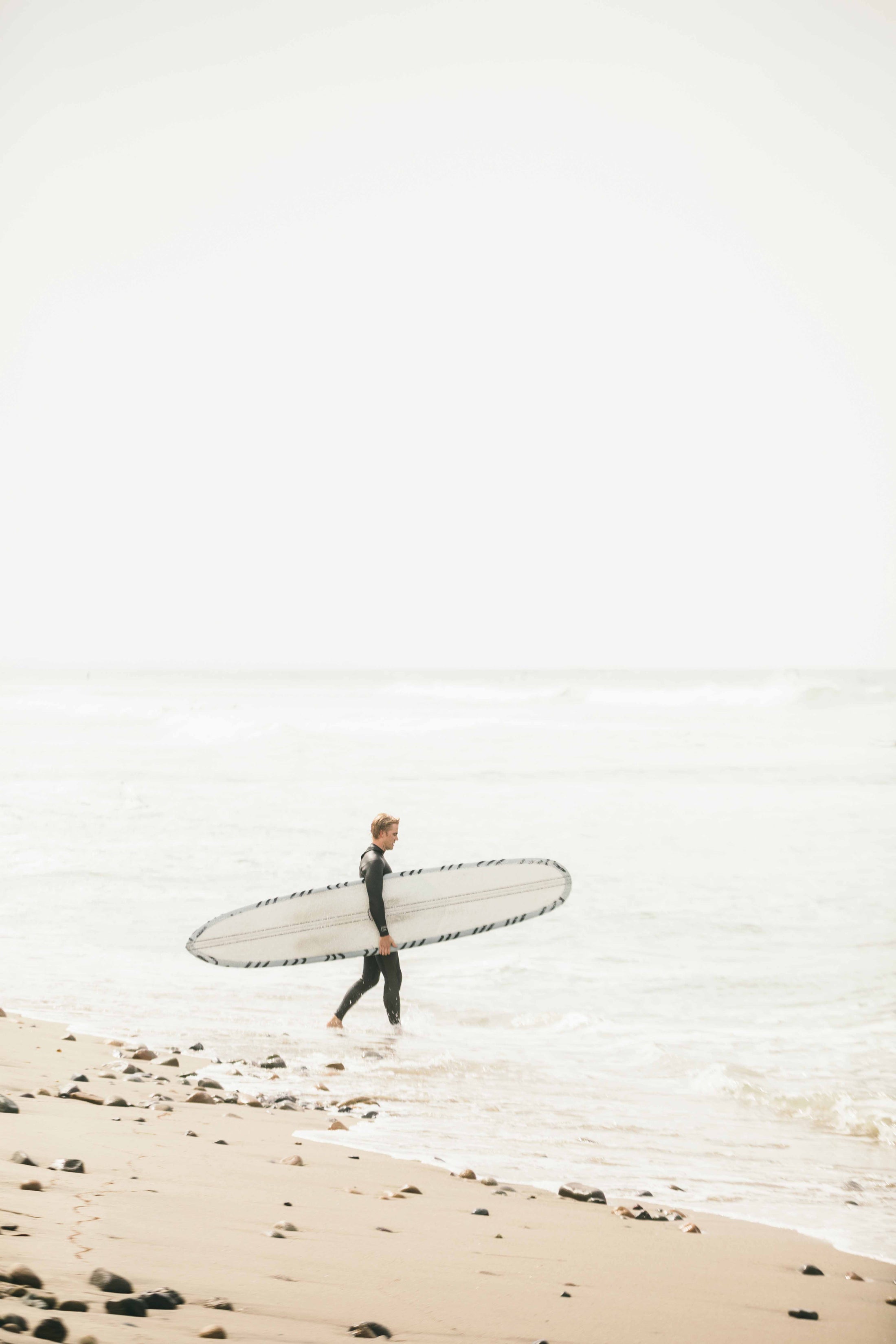 Man in wetsuit carrying long surfboard along rocky beach shore