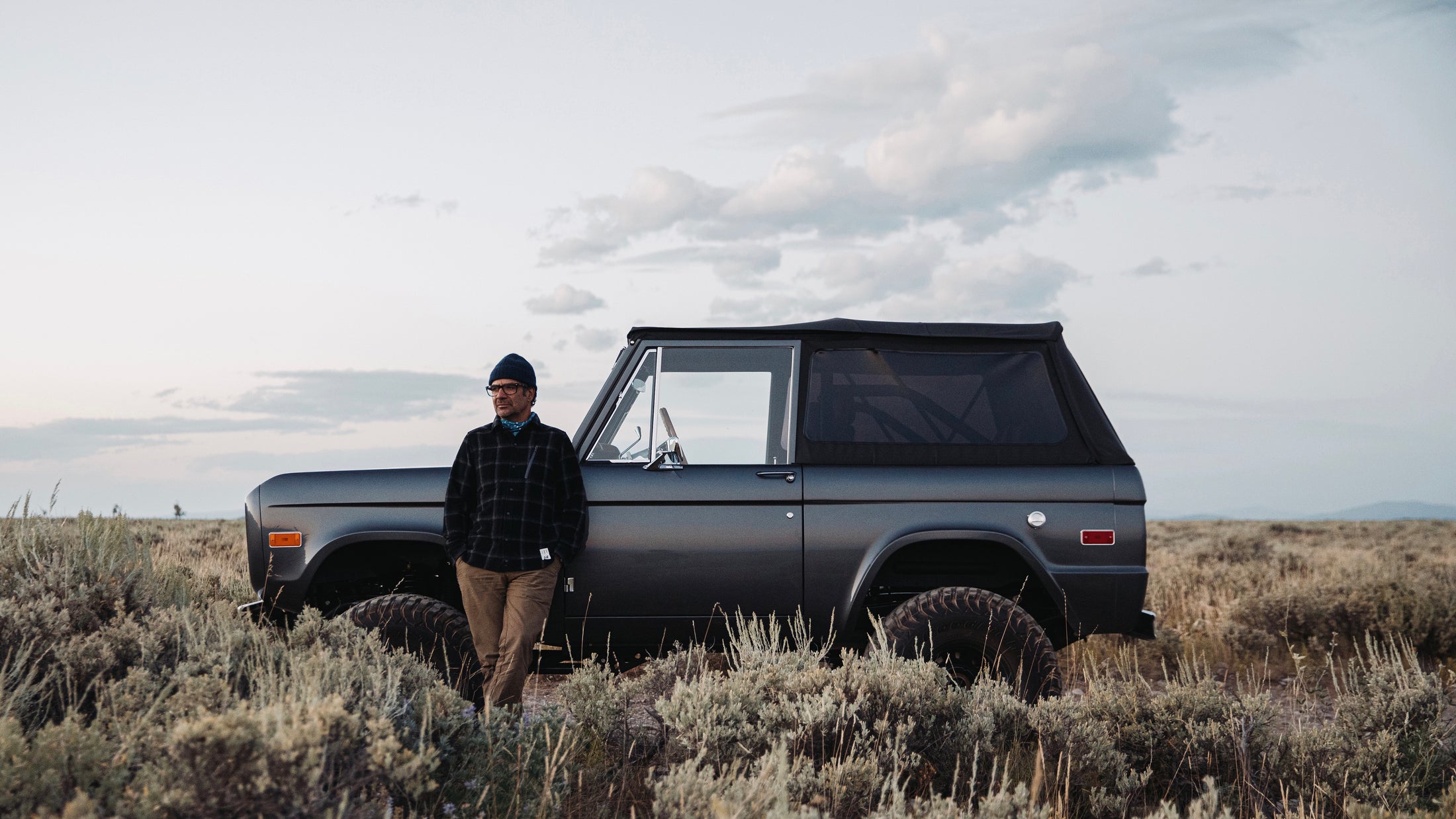 Classic Ford Bronco in gray with man wearing hat and glasses leaning on vehicle in shrub-filled landscape