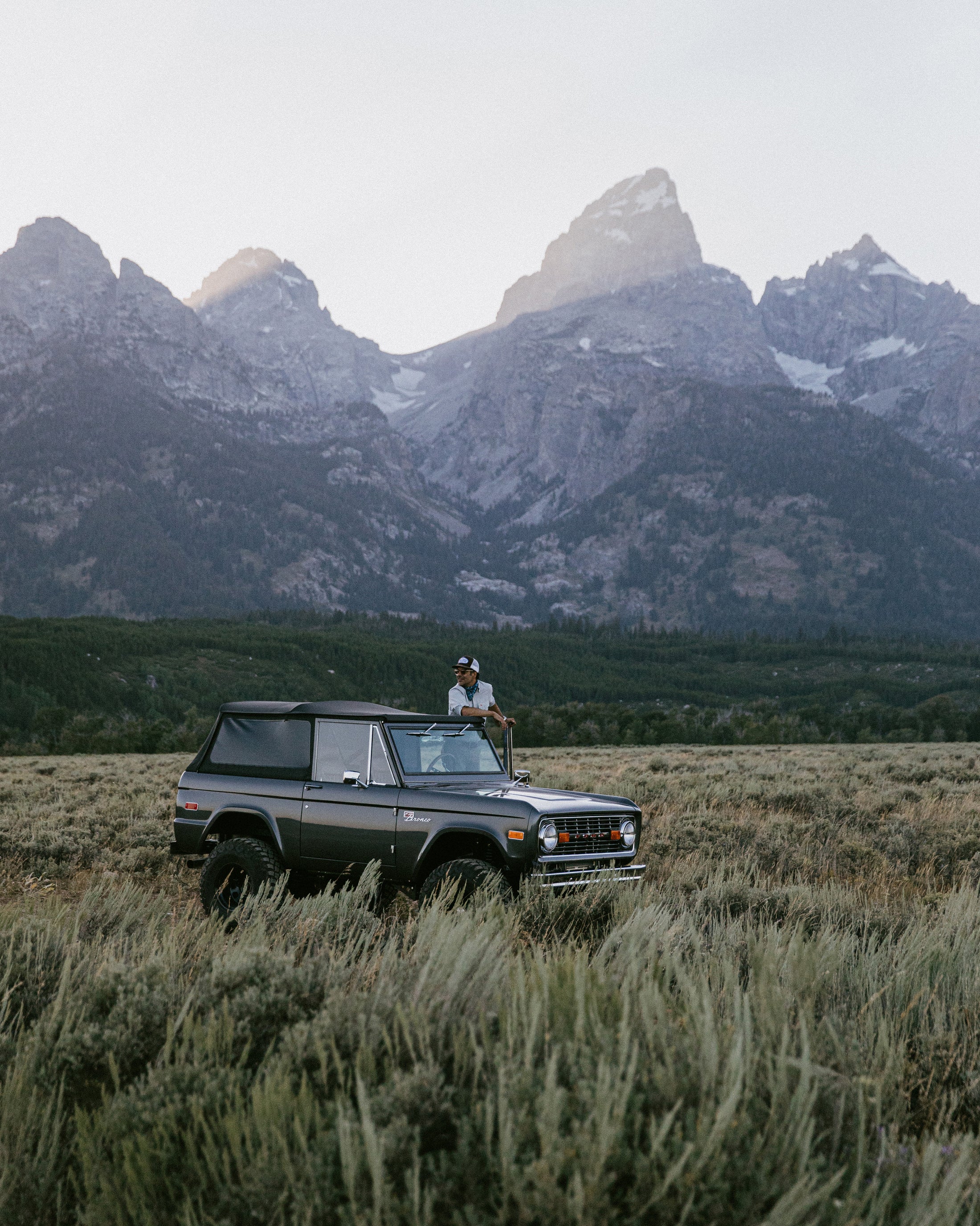 Classic Ford Bronco parked in grassy field with person standing through sunroof with mountainous backdrop