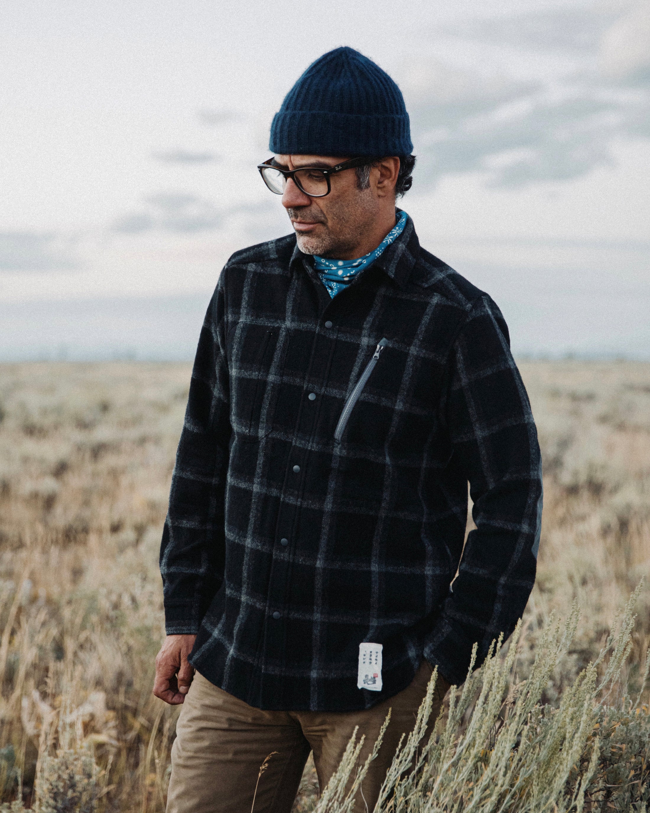 Man wearing blue beanie and glasses looking down, standing in tall dry grass