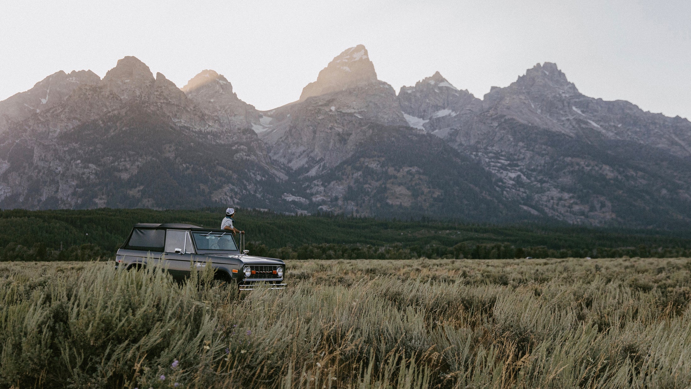 Classic Ford Bronco parked in grassy field with person standing through sunroof and mountains in background