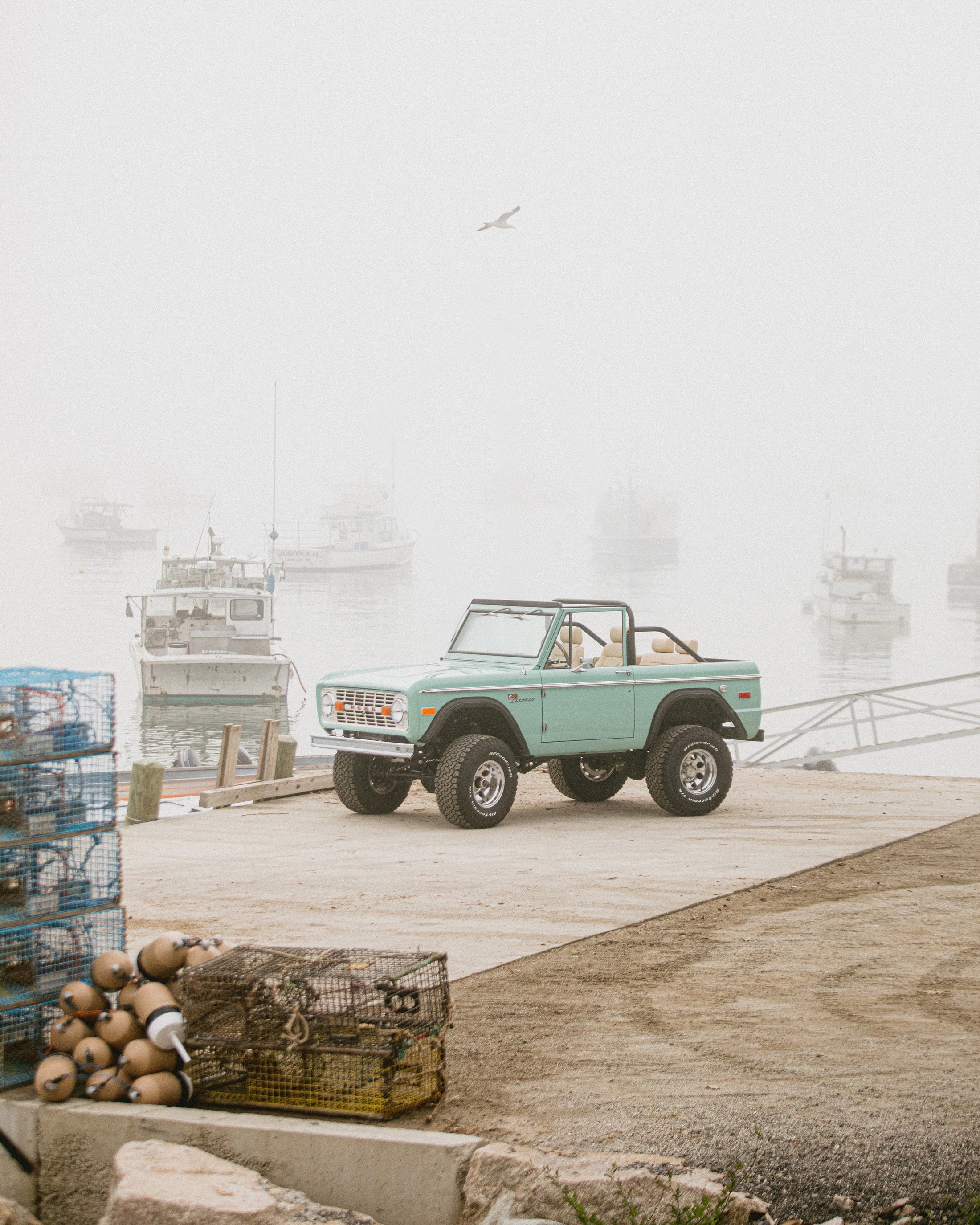 Classic Ford Bronco in light green parked on a concrete dock by boats in foggy harbor.