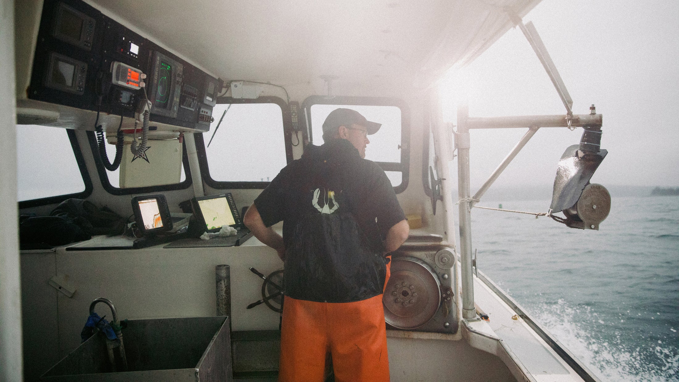 Man in orange bib pants and black shirt operating a boat's wheel inside a cabin