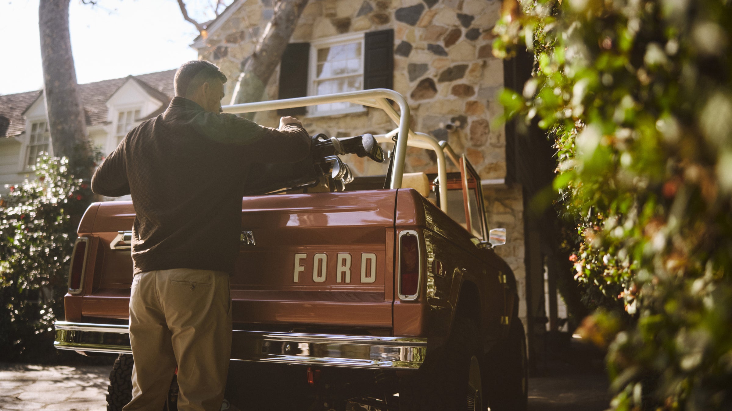 Man standing by the tailgate of a Classic Ford Bronco with the top down near a stone house