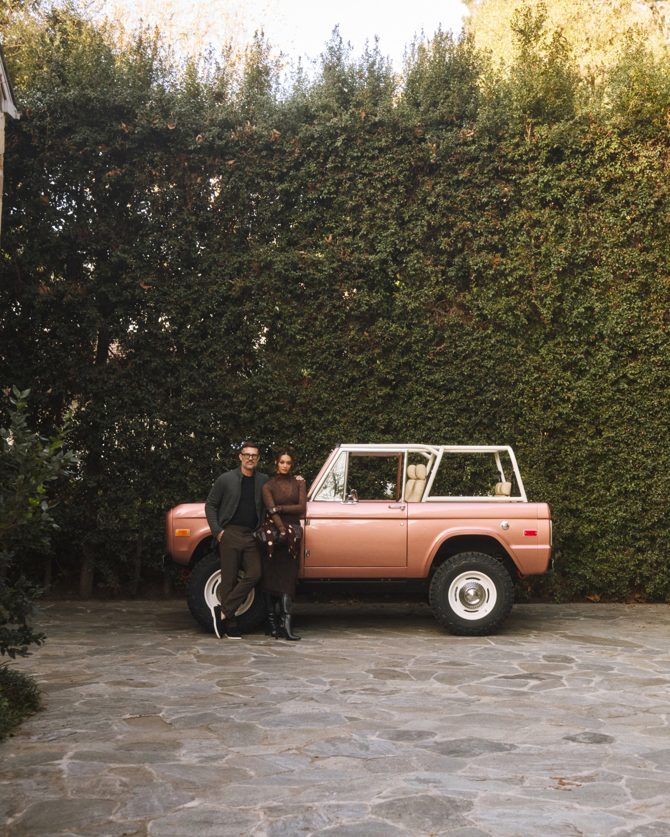 Man and woman standing beside a red Classic Ford Bronco with white wheels in front of a leafy hedge