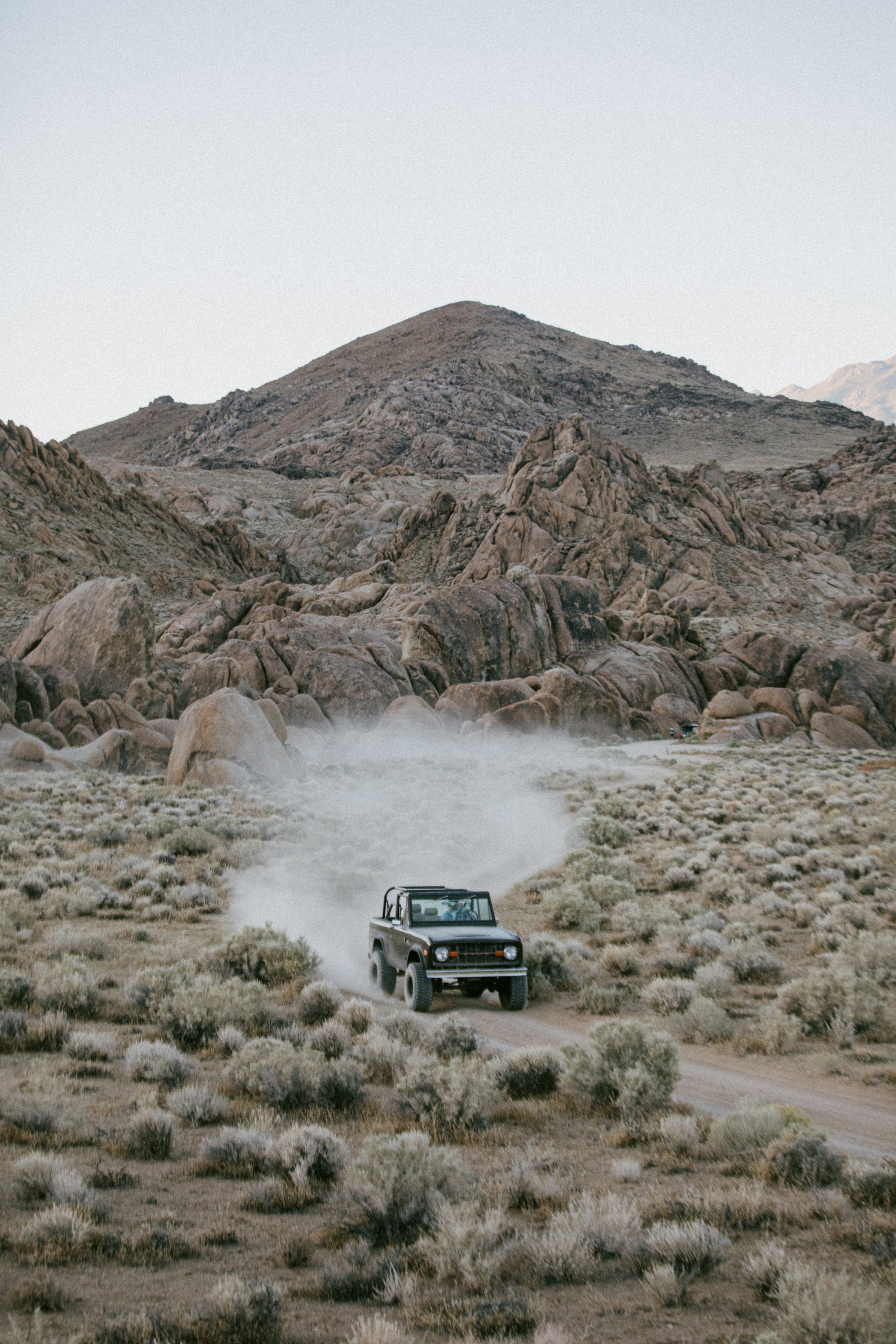 Classic Ford Bronco moving through desert vegetation toward rugged rocky terrain under dim light