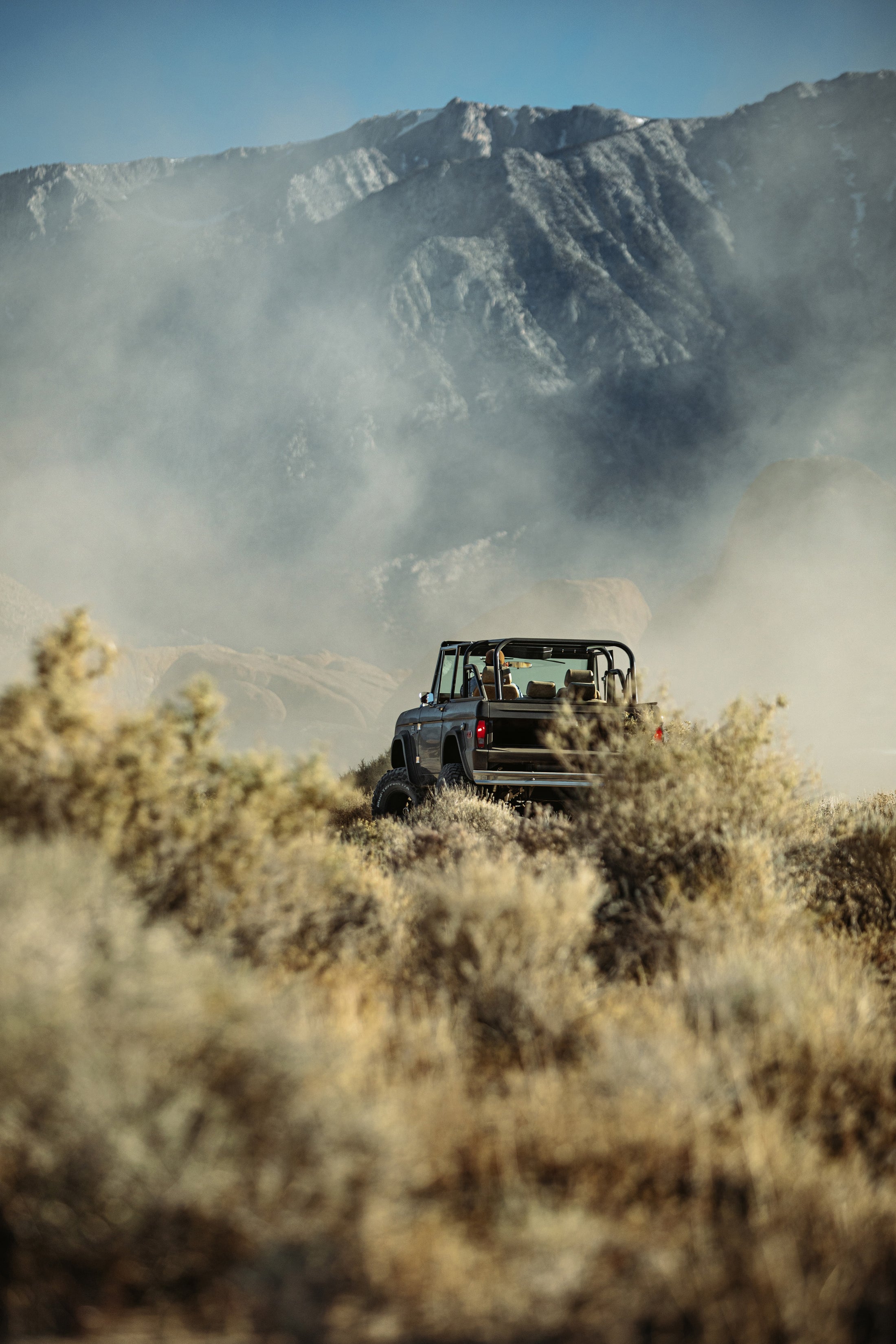 Classic Ford Bronco driving away on a trail through dry brush with mountain ridges in the distance