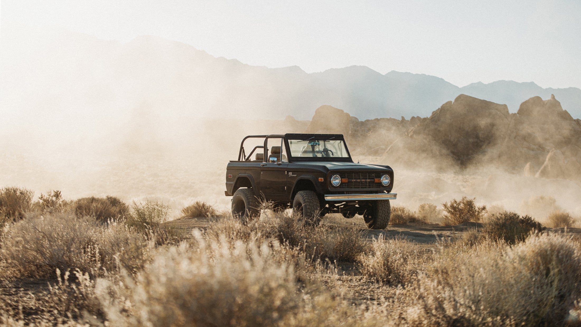 Classic Ford Bronco kicking up dust while driving through dry brush with rocky hills in background