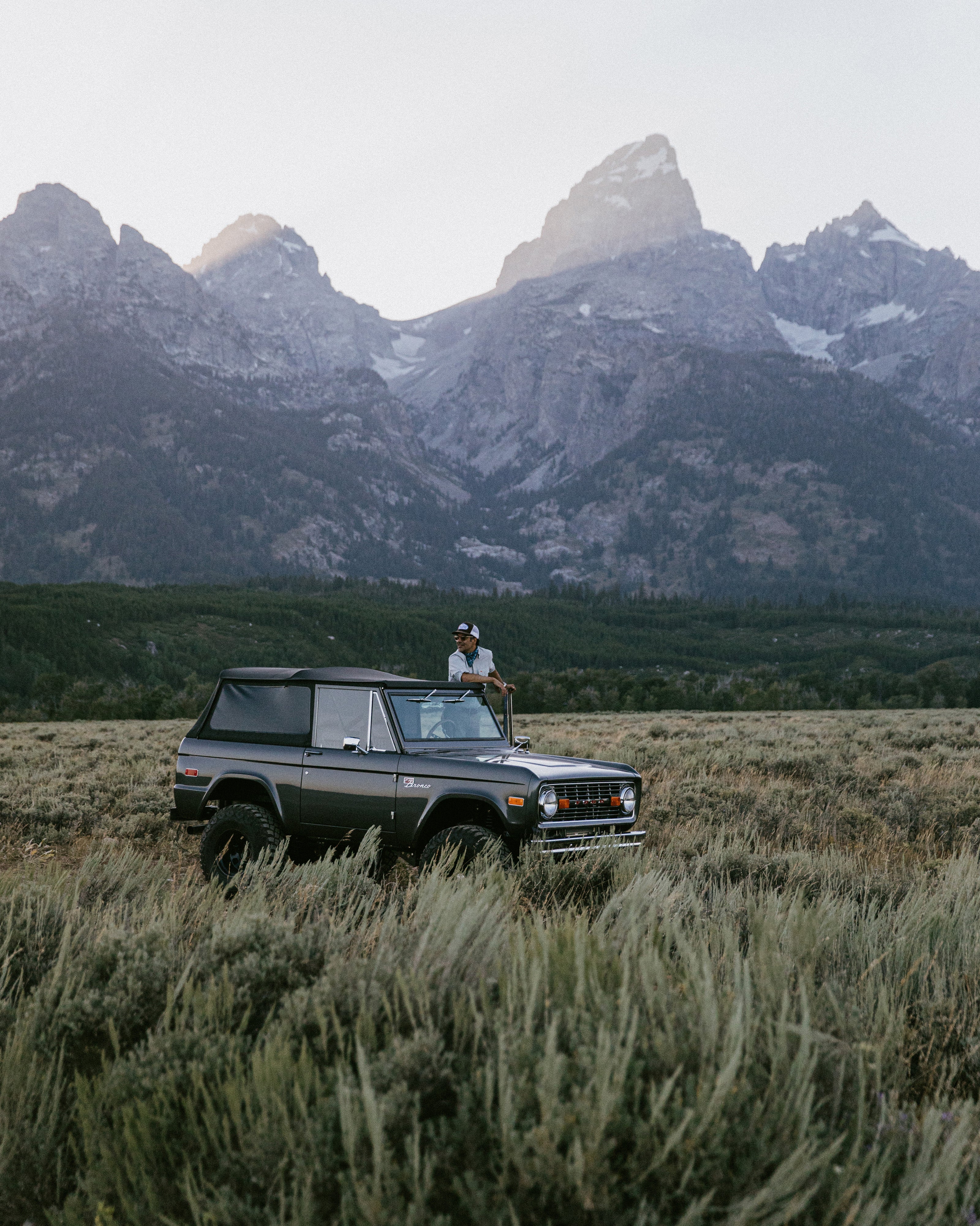 Classic Ford Bronco parked in a field with mountains in the background and person leaning on roof