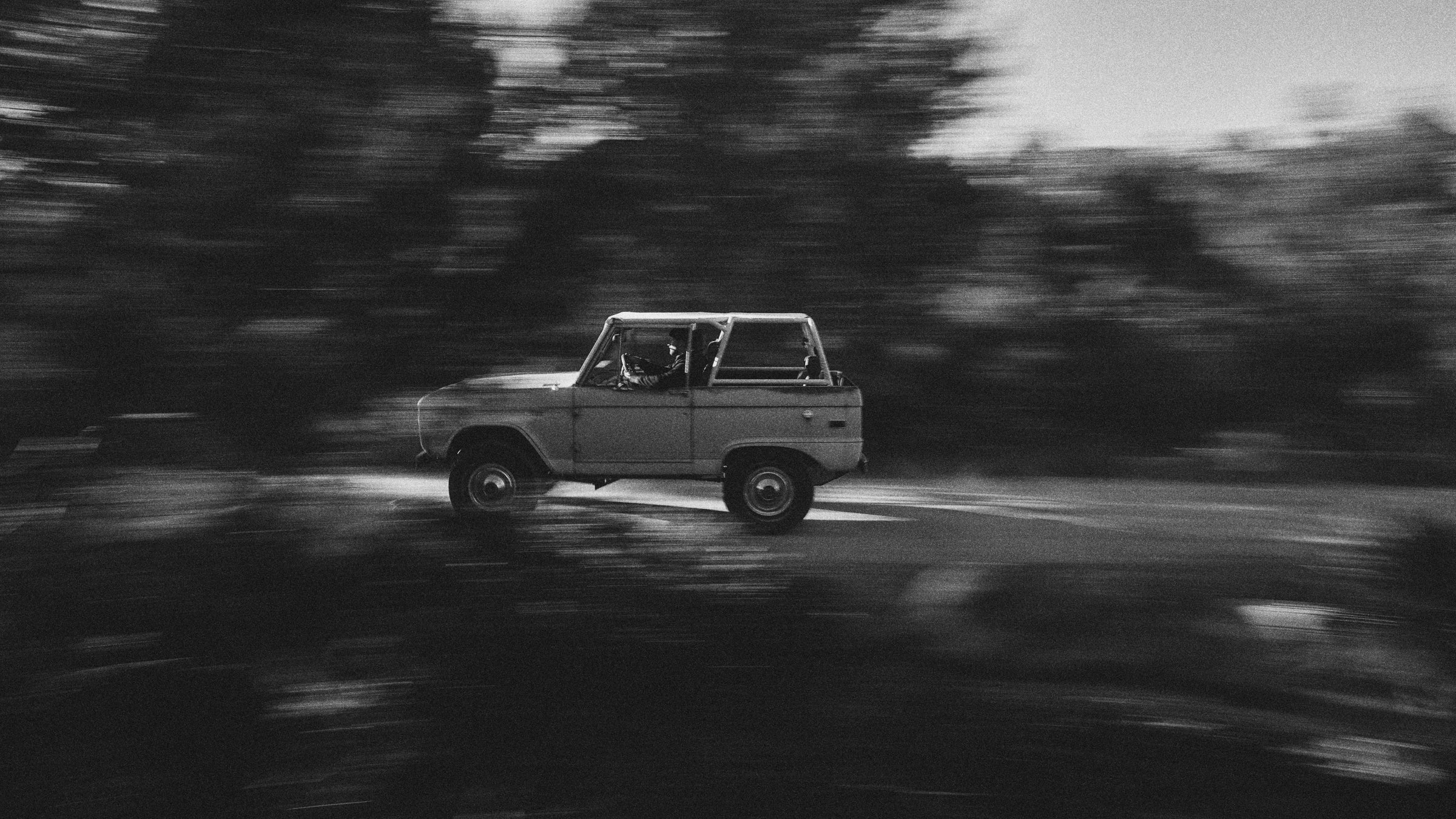 Black and white side view of a Classic Ford Bronco driving on a road surrounded by trees